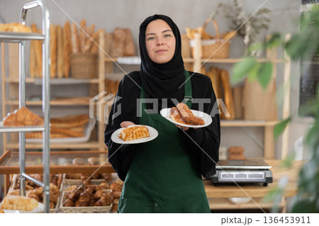 European Muslim girl employee of bakery stands with sandwiches in disposable plates European Muslim girl employee of bakery stands with sandwiches in disposable plates 136453911