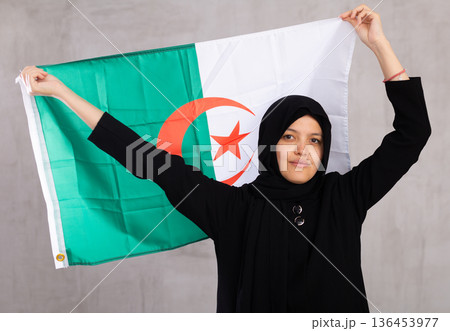 Young Muslim woman in black hijab holds unfurled flag of Algeria. 136453977