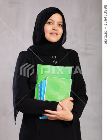 Portrait of young woman in hijab holding stack of notebooks Portrait of young woman in hijab holding stack of notebooks 136453998