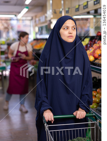 Muslim woman in dark blue hijab stands in grocery department of supermarket with grocery basket Muslim woman in dark blue hijab stands in grocery department of supermarket with grocery basket 136454019