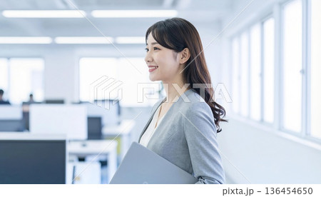ノートパソコンを持つ若い女性社員・明るい現代オフィスのビジネスシーン 136454650