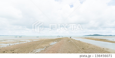 Wide shot of a coastal sandbar during low tide with traditional fishing boats in the distance. Minimalist seascape featuring a bright cloudy sky and calm ocean water at a tropical beach. 136456071