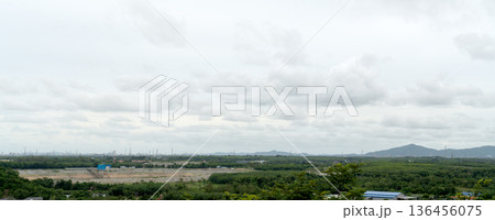 High-angle panoramic view of an industrial zone with factories on the horizon, surrounded by green forests, a dam reservoir, and mountains under a cloudy sky. Wide environmental landscape photo. 136456075