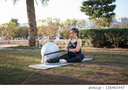 Pregnant Woman Doing Outdoor Stretching With Resistance Band And Exercise Ball On Mat Pregnant Woman Doing Outdoor Stretching With Resistance Band And Exercise Ball On Mat 136465447