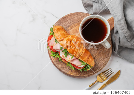 Croissant sandwich with ham, mozzarella cheese, tomato, arugula on a wooden board and cup of coffee Croissant sandwich with ham, mozzarella cheese, tomato, arugula on a wooden board and cup of coffee 136467403