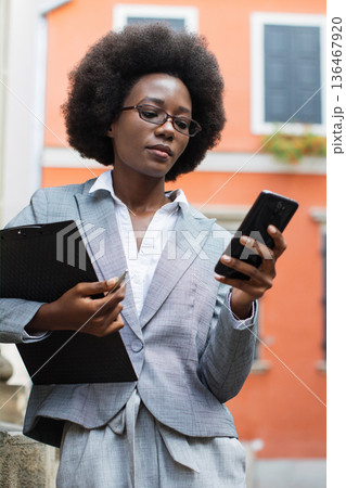 A professional Black woman in a suit holds a clipboard and a smartphone while looking at the device outdoors with an orange building in the background A professional Black woman in a suit holds a clipboard and a smartphone while looking at the device outdoors with an orange building in the background 136467920