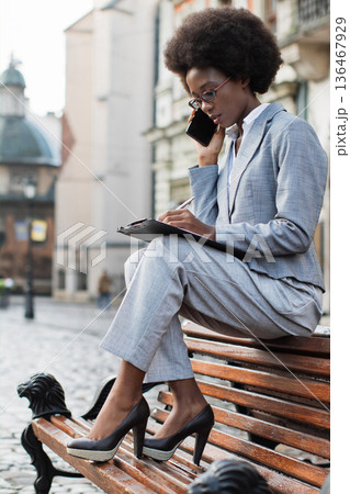 A professional Black woman in a suit sits on a park bench outdoors, talking on her phone while taking notes on a clipboard 136467929