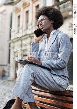 A professional Black woman in a suit sits on a bench outdoors, talking on her phone while taking notes on a clipboard A professional Black woman in a suit sits on a bench outdoors, talking on her phone while taking notes on a clipboard 136467933