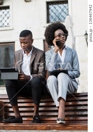 A man and a woman in business attire sit on a wooden bench outdoors, engaged with their digital devices and phones A man and a woman in business attire sit on a wooden bench outdoors, engaged with their digital devices and phones 136467942