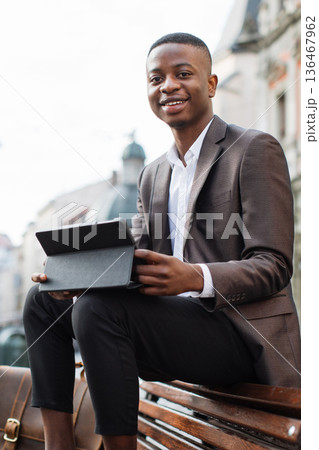 A smiling young Black man in a suit sits on a park bench holding a tablet device with a leather satchel beside him A smiling young Black man in a suit sits on a park bench holding a tablet device with a leather satchel beside him 136467962