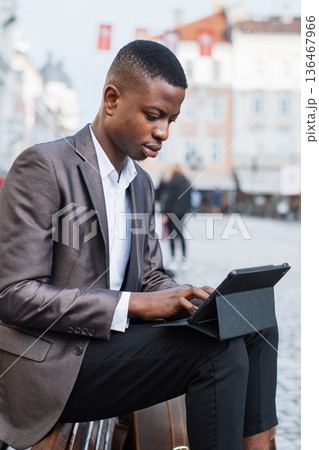 A young Black man in a suit jacket and white shirt sits outdoors on a bench, focused on typing on a tablet device A young Black man in a suit jacket and white shirt sits outdoors on a bench, focused on typing on a tablet device 136467966