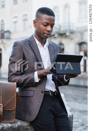 A young professional man in a suit uses a tablet device while standing outside in an urban environment A young professional man in a suit uses a tablet device while standing outside in an urban environment 136467970