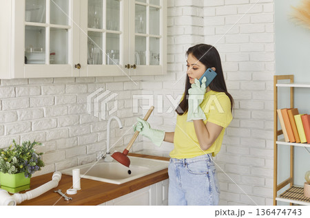 Worried young woman with plunger standing in kitchen and calling plumber to fix clogged sink. 136474743