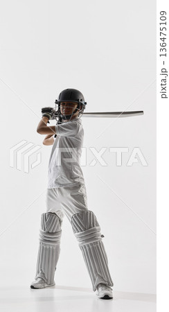 Young cricket player holding bat over shoulder in protective gear on white background. Young cricket player holding bat over shoulder in protective gear on white background. 136475109