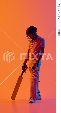 Young cricket batsman standing with bat grounded on vibrant orange gradient background. Young cricket batsman standing with bat grounded on vibrant orange gradient background. 136475213