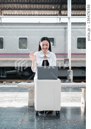 Young woman at train station using laptop on suitcase, smiling and waving, representing modern travel and communication. Young woman at train station using laptop on suitcase, smiling and waving, representing modern travel and communication. 136476386
