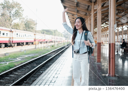 Smiling woman at train station platform, waving with camera and backpack, ready for travel adventure. Smiling woman at train station platform, waving with camera and backpack, ready for travel adventure. 136476400