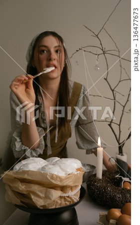 In lace headscarf and khaki apron, she smiles decorating Easter cake with white icing, creating warm festive kitchen atmosphere. In lace headscarf and khaki apron, she smiles decorating Easter cake with white icing, creating warm festive kitchen atmosphere. 136477673