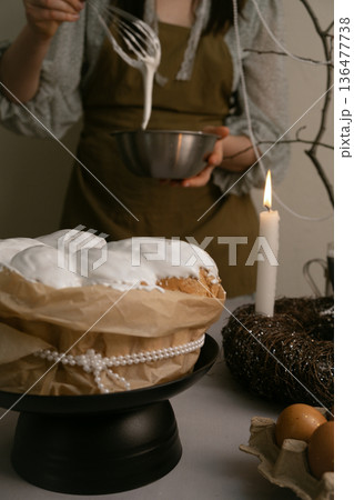 Cozy Easter moment in kitchen, she carefully spreads icing on cake, with eggs, candles, wreath on table, beaded branch in background. Cozy Easter moment in kitchen, she carefully spreads icing on cake, with eggs, candles, wreath on table, beaded branch in background. 136477738