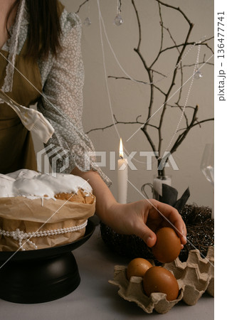 Woman in home clothes carefully decorates Easter cake with white icing, eggs, candles, wreath, and beaded branch creating festive atmosphere. 136477741