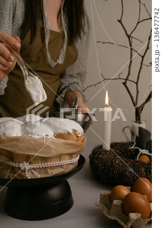 Preparing for holiday, woman spreads white icing on Easter cake with brush, table decorated with eggs, candles, wreath, and beaded branch. Preparing for holiday, woman spreads white icing on Easter cake with brush, table decorated with eggs, candles, wreath, and beaded branch. 136477742