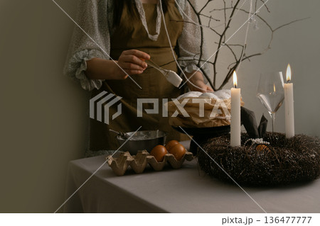 Woman carefully brushes white icing on Easter cake, with eggs, candles, and twig wreath on table, decorated branch in background. 136477777