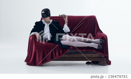 Woman in black jacket and ruffled blouse on red velvet sofa holding opera glasses. 136479781