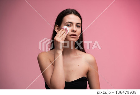 Young woman applying micellar water with a cotton pad to clean her face and remove makeup, focusing on daily skincare and beauty treatment for healthy, clear skin against a pink background Young woman applying micellar water with a cotton pad to clean her face and remove makeup, focusing on daily skincare and beauty treatment for healthy, clear skin against a pink background 136479939