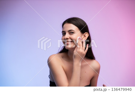 Smiling woman cleansing skin, applying facial toner with a cotton pad for daily skincare routine, focusing on health, beauty, and cosmetic treatment against a vibrant background 136479940