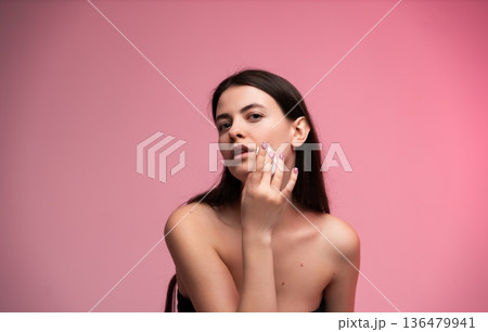Young woman applying skincare product to her face, using cotton pad for cleansing, focusing on beauty and health with fresh, glowing skin on pink background 136479941