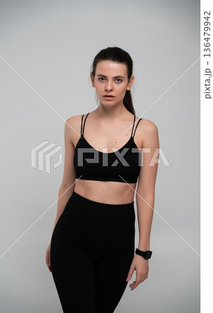 Young athletic woman in black activewear and fitness tracker standing in studio, looking at camera, representing health, wellness, and dedicated workout lifestyle 136479942