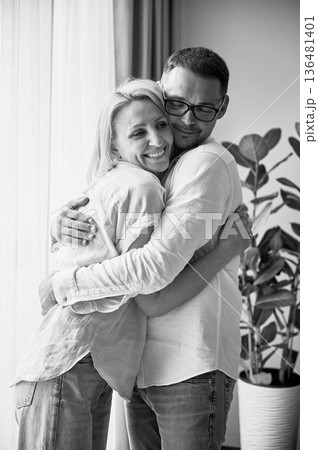 Happy couple hugging on light background at home. Pretty woman smiling and hugging her husband. Sweet couple in casual clothes. Black and white image. Happy couple hugging on light background at home. Pretty woman smiling and hugging her husband. Sweet couple in casual clothes. Black and white image. 136481401