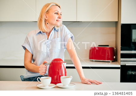 Pretty female making tea for two. Young woman holding red kettle and brewing tea. Beautiful woman looking away and smiling. Pretty female making tea for two. Young woman holding red kettle and brewing tea. Beautiful woman looking away and smiling. 136481405