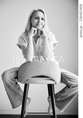 Pretty female relaxing on bar stool. Portrait of woman with blonde hair smiling straight to camera. Beautiful woman sitting on bar stool in her apartment. Black and white image. 136481408