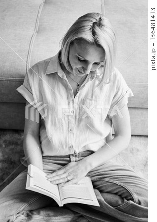 Gentle female sitting and reading next to sofa. Woman reading interesting book. Pretty woman relaxing on ream carpet. Black and white image. 136481413