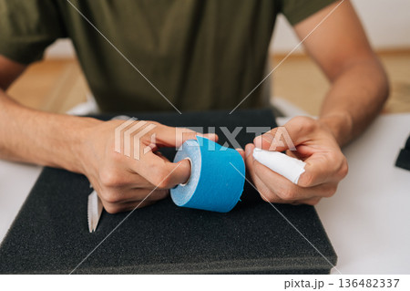 Close-up cropped shot of man holding roll of kinesiology tape on table, preparing to apply for muscle, joint or tendon pain relief, representing self-care solution for aches and injuries. 136482337