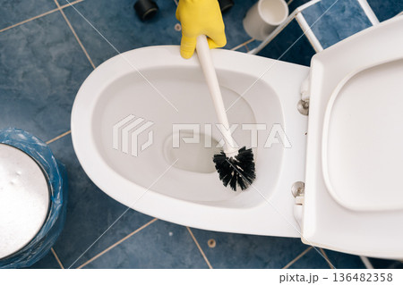 Close-up of hand in yellow protective glove cleaning ceramic toilet bowl with brush, maintaining cleanliness and hygiene standards in residential or commercial washroom environment. Close-up of hand in yellow protective glove cleaning ceramic toilet bowl with brush, maintaining cleanliness and hygiene standards in residential or commercial washroom environment. 136482358