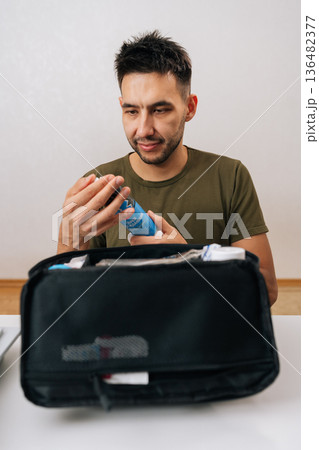 Vertical portrait of young man reaching for medication from first aid kit for self-care, health treatment and emergency healing at home sitting at table on isolated white background. Vertical portrait of young man reaching for medication from first aid kit for self-care, health treatment and emergency healing at home sitting at table on isolated white background. 136482377