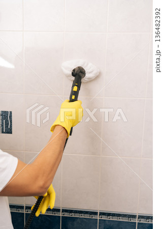 Close-up of housekeeper in yellow gloves using steam cleaner to deep-clean and sanitize white bathroom tiles, removing dirt and germs for a hygienic, comfortable home environment. Close-up of housekeeper in yellow gloves using steam cleaner to deep-clean and sanitize white bathroom tiles, removing dirt and germs for a hygienic, comfortable home environment. 136482392