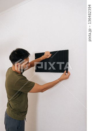 Vertical back view of man fixing applying sound absorbing foam panels to wall, working on acoustic treatment for home studio or office, making soundproofing adjustments and managing noise reduction. Vertical back view of man fixing applying sound absorbing foam panels to wall, working on acoustic treatment for home studio or office, making soundproofing adjustments and managing noise reduction. 136482394