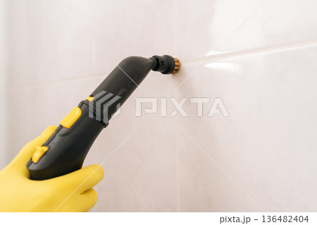 Close-up of hand in yellow protective glove using steam cleaner with brush attachment to disinfect and clean grout lines between white tiles in clean bathroom. Concept of cleanliness and housekeeping. Close-up of hand in yellow protective glove using steam cleaner with brush attachment to disinfect and clean grout lines between white tiles in clean bathroom. Concept of cleanliness and housekeeping. 136482404