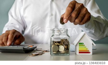 Businessman putting a coin into a jar for home savings and energy efficiency planning. Strategic concept for sustainable housing investment, green energy costs, mortgage calculation, and real estate 136483810