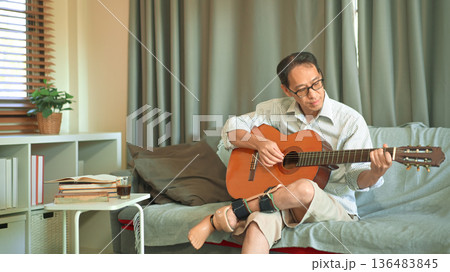 Man playing acoustic guitar on a sofa at home, wearing a prosthetic leg, relaxed leisure time and independent adaptive lifestyle. Man playing acoustic guitar on a sofa at home, wearing a prosthetic leg, relaxed leisure time and independent adaptive lifestyle. 136483845
