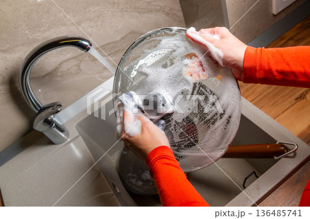 A close-up with localized lighting. The hands of a Caucasian woman washing dishes in a home kitchen late at night in the dark. A close-up with localized lighting. The hands of a Caucasian woman washing dishes in a home kitchen late at night in the dark. 136485741