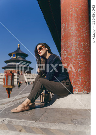 brunette girl sitting on the steps by temple of heaven in China. Brunette girl enjoying chinese architecture while sitting on the steps and relaxing. Caucasian tourist taking a break from exploring brunette girl sitting on the steps by temple of heaven in China. Brunette girl enjoying chinese architecture while sitting on the steps and relaxing. Caucasian tourist taking a break from exploring 136486042