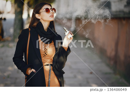 stylish girl smoking an e-cigarette as she is walking through the city stylish girl smoking an e-cigarette as she is walking through the city 136486049