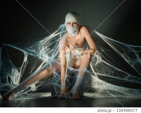 Woman in mummy costume with dramatic eye makeup sitting trapped in artificial spiderwebs in a dark, spooky setting for Halloween. 136486077