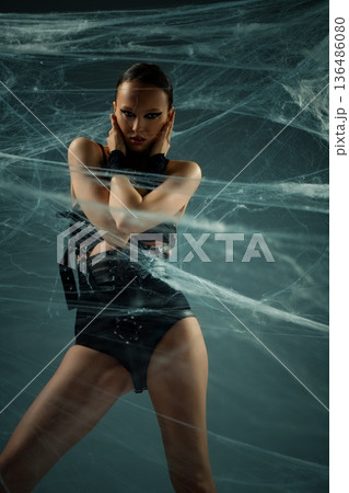 Intense woman in black gothic fashion outfit posing behind a dense spiderweb. 136486080