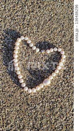 Pearl necklace arranged to form a heart shape on sandy beach during sunny day with natural light casting shadows Pearl necklace arranged to form a heart shape on sandy beach during sunny day with natural light casting shadows 136486296