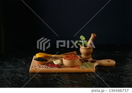 Professional still life photography showcasing assorted culinary spices and fresh herbs arranged on wooden cutting board featuring mortar and pestle on dark marble against black background 136486737
